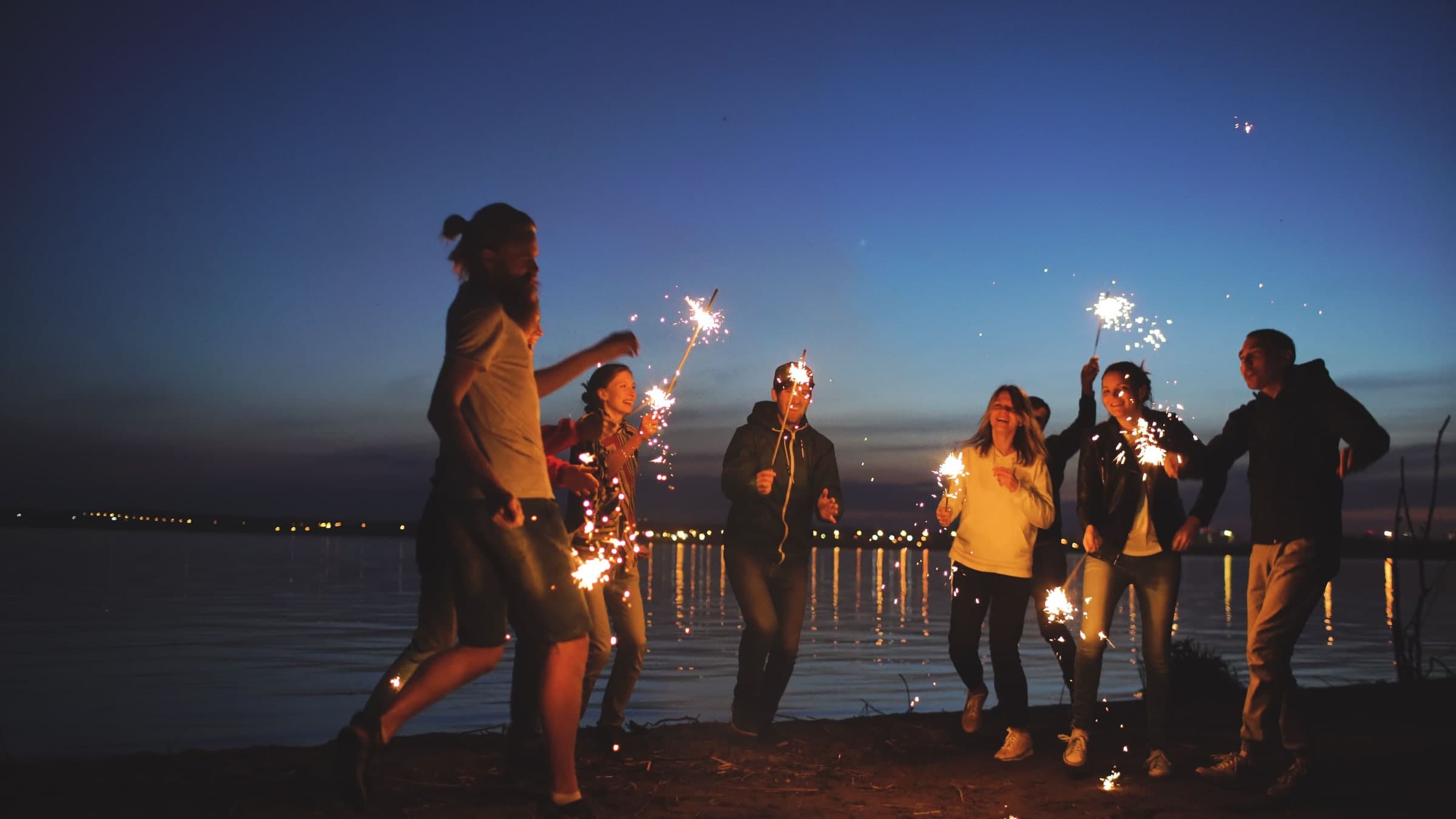Friends laughing and dancing with sparklers on a beach at dusk